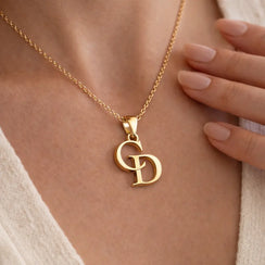 A close-up of a gold chain necklace with a monogram pendant featuring the letters 'G' and 'D', worn on a model's neck with a hand resting near the jewelry.