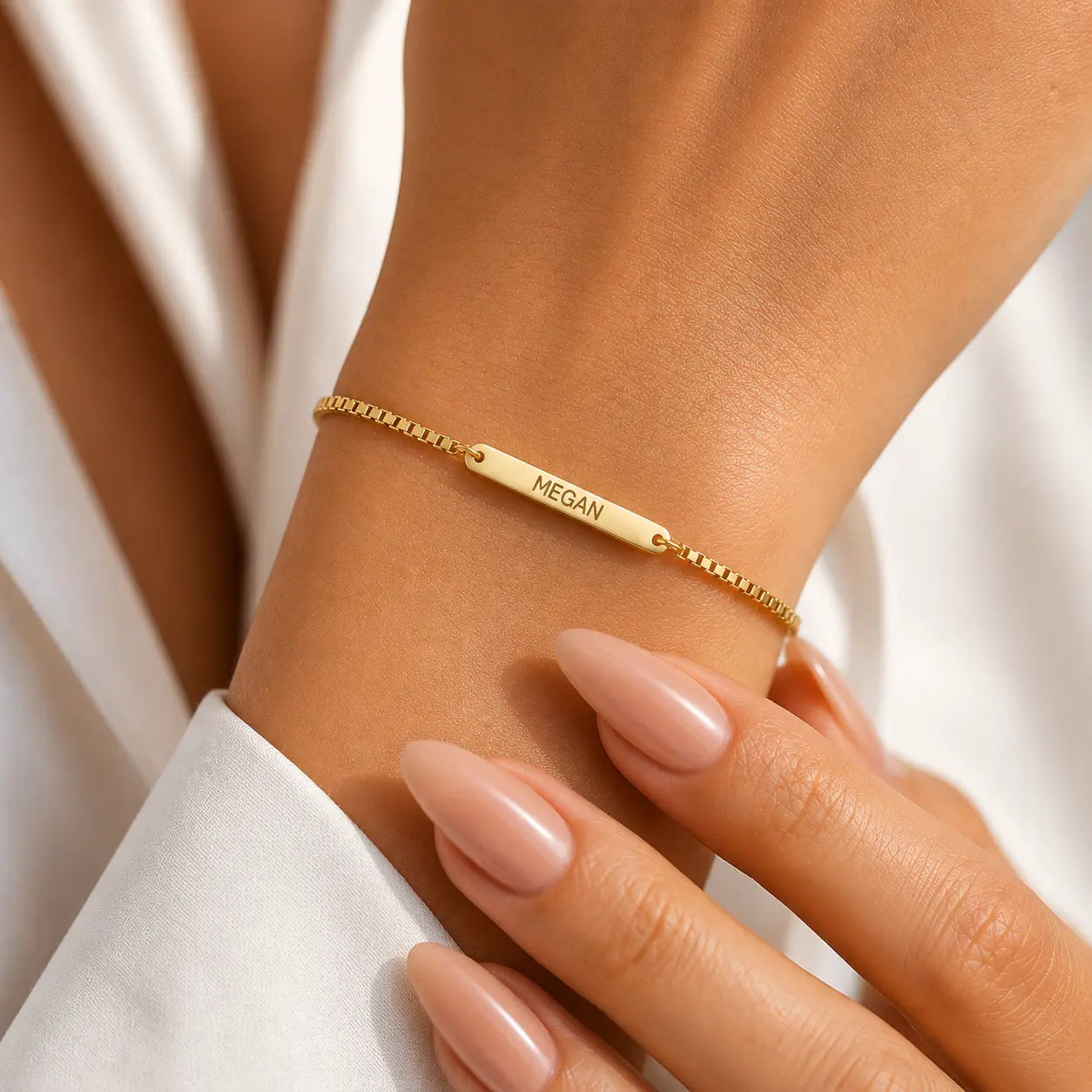 A delicate gold chain bracelet with a central bar engraved with the name 'Megan', shown worn on a person's wrist with manicured nails and a white sleeve visible.