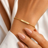 A delicate gold chain bracelet with a central bar engraved with the name 'Megan', shown worn on a person's wrist with manicured nails and a white sleeve visible.
