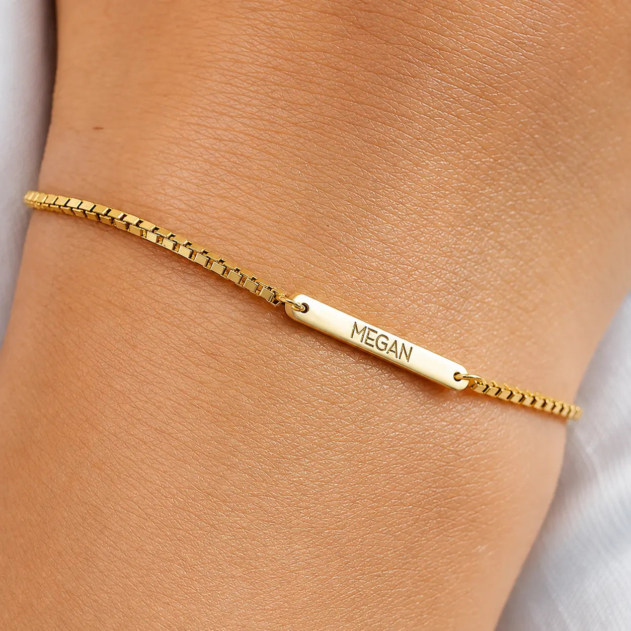 A gold chain bracelet worn on a wrist, featuring a polished bar engraved with the name 'MEGAN'.