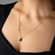 A close-up of a model wearing a delicate gold necklace with a script nameplate and a teardrop-shaped green gemstone pendant.