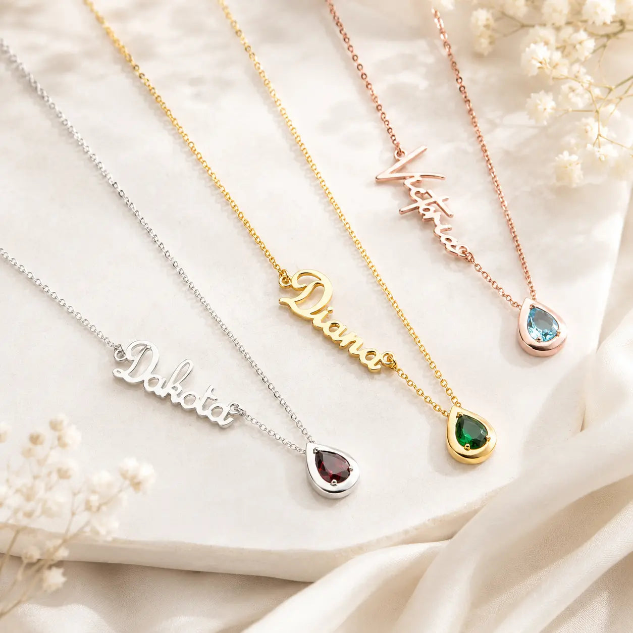 Four personalized name necklaces with teardrop gemstone pendants are arranged diagonally on a draped cream fabric background with small white flowers. The necklaces are in silver, yellow gold, rose gold, and a pink gold tone, with pendants reading Dakota, Diana, Victoria, and Kate.