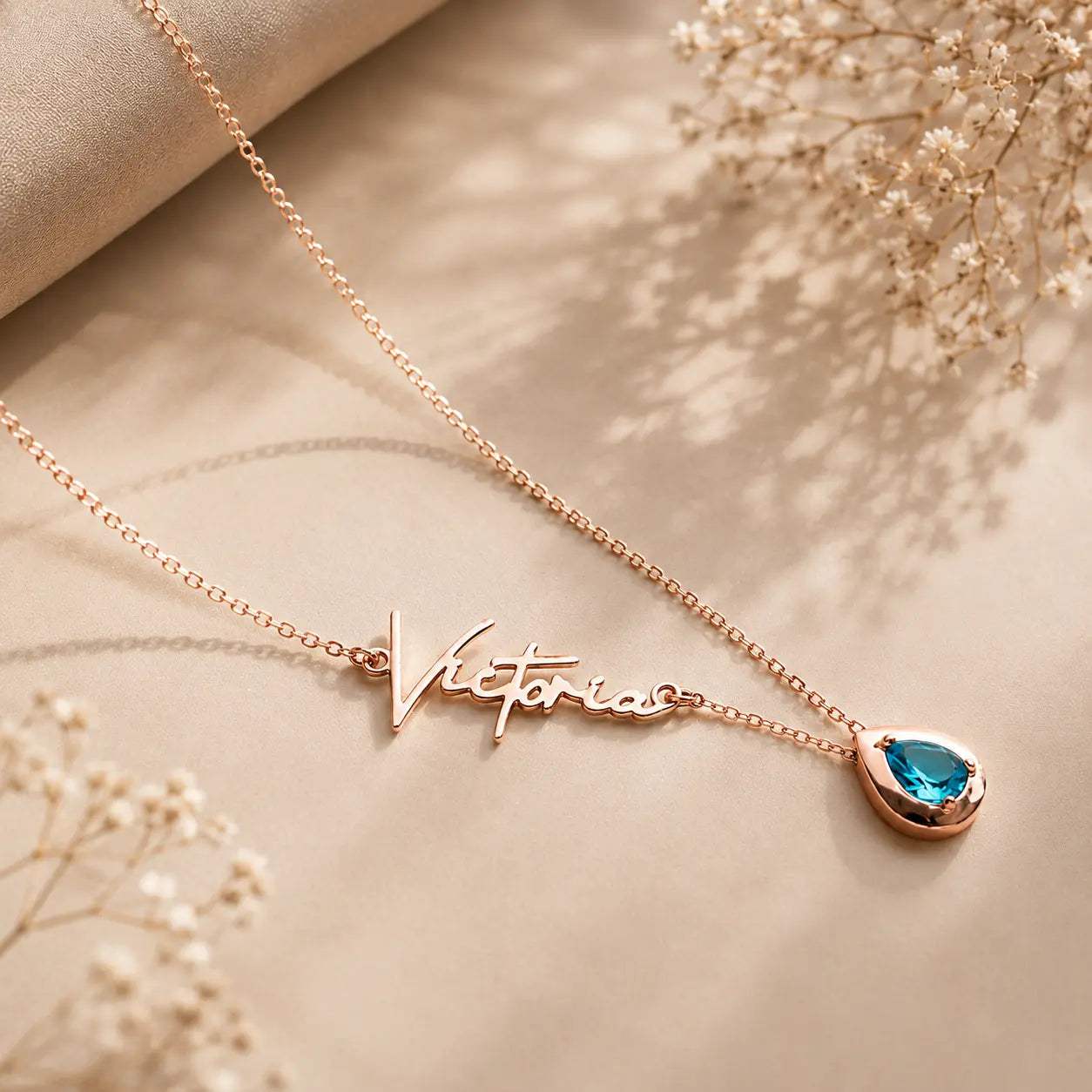 A rose gold script name necklace reading 'Victoria' with a teardrop-shaped blue gemstone pendant, styled on a beige fabric background with dried baby's breath flowers.