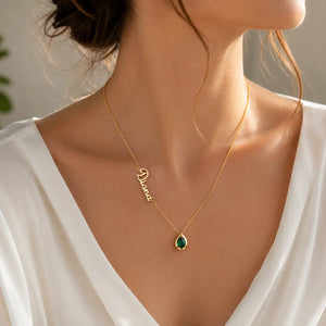 A close-up lifestyle shot of a model wearing a delicate gold name necklace with a green teardrop gemstone pendant and a cursive nameplate spelling 'Diane'.
