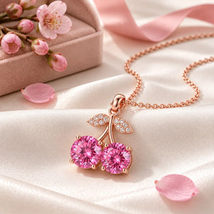 Rose gold cherry pendant necklace featuring two round pink gemstones in the shape of cherries, with leaf details paved with small clear stones, shown on a white background with a reflection.