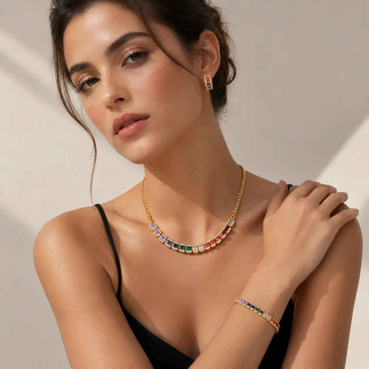 A woman models a gold jewelry set featuring a multi-colored rectangular-stone necklace and matching stud earring and bracelet.