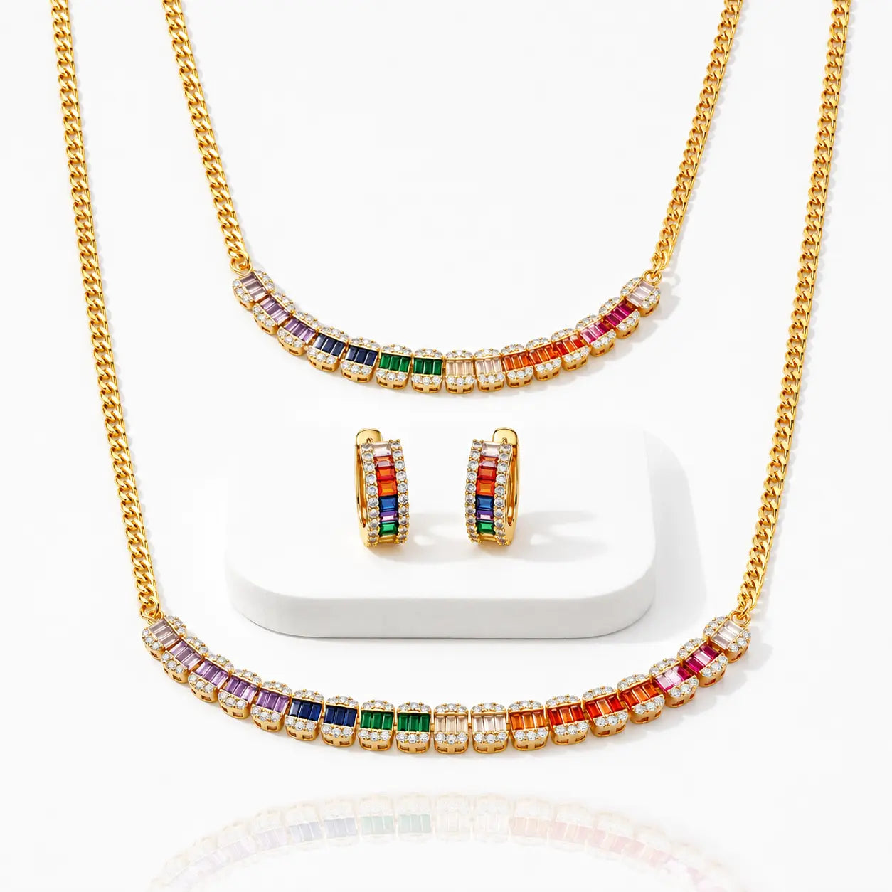 Gold jewelry set with a necklace, matching earrings, and a smaller necklace displayed on a white stand. The pieces feature colorful gemstone accents in purple, blue, green, orange, and red, set with clear stones on a white background.