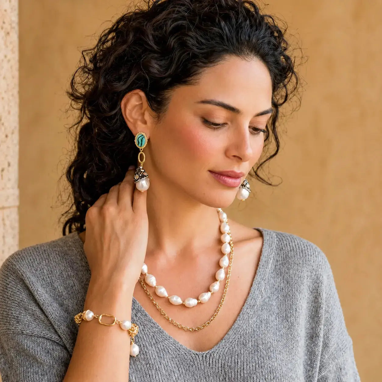A woman with curly dark hair models a matching jewelry set featuring a double-strand pearl necklace, a gold chain necklace, a gold bracelet with pearl accents, and matching drop earrings with turquoise and gold details, set against a warm beige background.