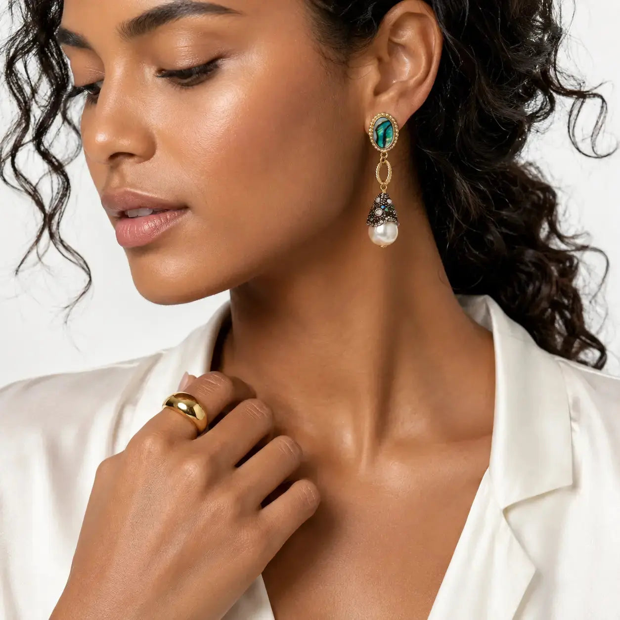 A close-up of a woman wearing a gold drop earring with a green oval stone, silver filigree, and a white pearl, paired with a gold ring on her finger.
