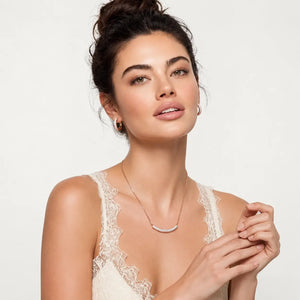 A woman wearing a delicate silver crescent moon necklace with a small clear gemstone, styled with matching gold hoop earrings and a cream lace camisole against a light gray background.