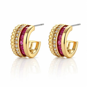 A pair of gold huggie hoop earrings featuring a central channel set with faceted pink gemstones and rows of small clear stones, shown on a white background with a soft reflection.