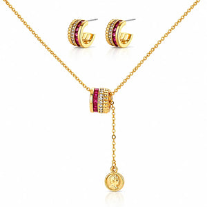 Gold jewelry set featuring a necklace with a cylindrical charm flanked by red gemstones and clear stones, matching hoop earrings, and a round pendant with an embossed profile, displayed on a white background.