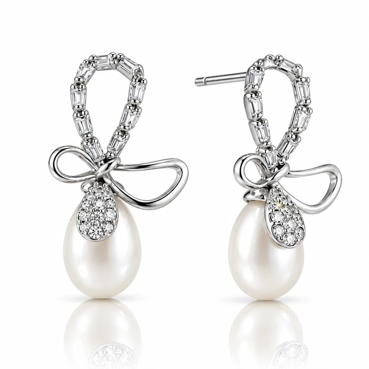A pair of silver drop earrings featuring a bow-shaped top set with baguette and round clear stones, a teardrop-shaped clear stone pavé setting, and a large white pearl dangling below, shown on a white background with a soft reflection.