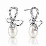 A pair of silver drop earrings featuring a bow-shaped top set with baguette and round clear stones, a teardrop-shaped clear stone pavé setting, and a large white pearl dangling below, shown on a white background with a soft reflection.