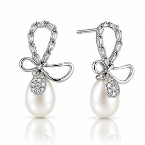 A pair of silver drop earrings featuring a bow-shaped top set with baguette and round clear stones, a teardrop-shaped clear stone pavé setting, and a large white pearl dangling below, shown on a white background with a soft reflection.