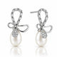 A pair of silver drop earrings featuring a bow-shaped top set with baguette and round clear stones, a teardrop-shaped clear stone pavé setting, and a large white pearl dangling below, shown on a white background with a soft reflection.