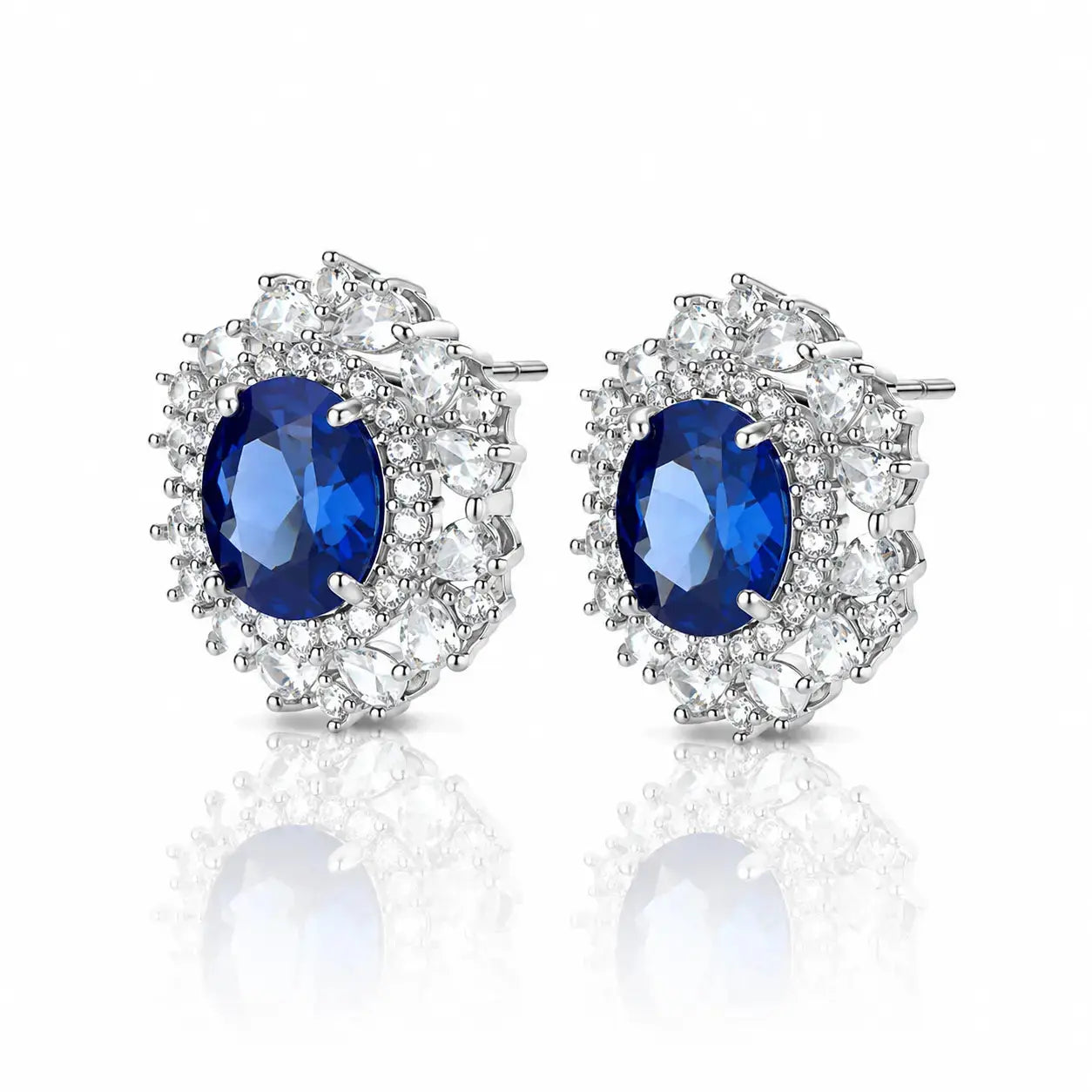 A pair of oval stud earrings featuring a central deep blue gemstone surrounded by a halo of clear round and marquise-cut stones in a silver-toned setting, shown on a white background with a soft reflection below.