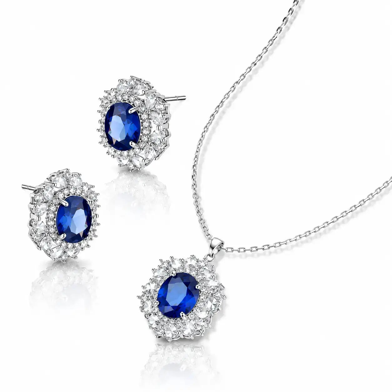 A jewelry set featuring a silver necklace and matching stud earrings, each adorned with a central oval-cut deep blue gemstone surrounded by a halo of small clear stones, displayed on a clean white background.