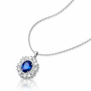 A silver chain necklace with a teardrop-shaped pendant featuring a central deep blue gemstone surrounded by a halo of small clear stones, shown on a white background with a soft reflection.
