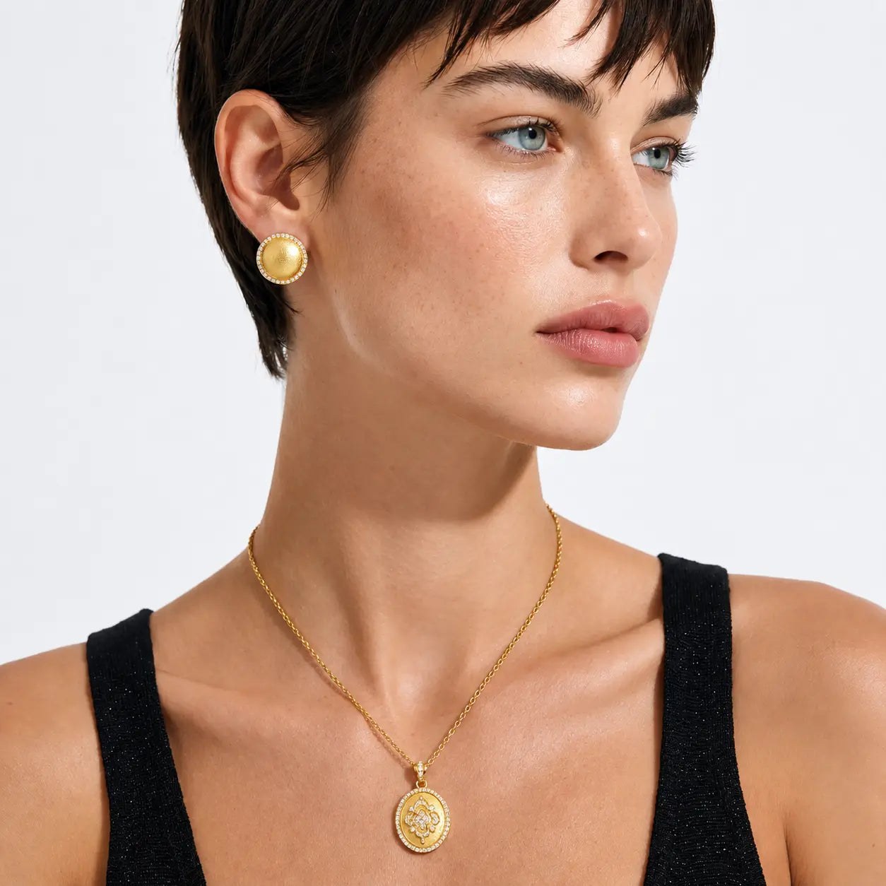A close-up of a model wearing a gold pendant necklace and matching gold stud earring against a clean white background.