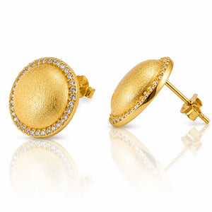A pair of gold oval stud earrings with a textured center and a border of small clear stones, shown on a white reflective background.