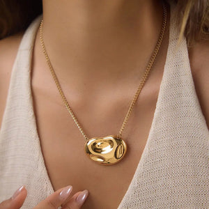 A close-up of a model wearing a gold chain necklace with a polished, organic-shaped gold pendant, styled with a beige knit top.