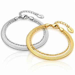 Two double-strand snake chain bracelets, one in silver and one in yellow gold, displayed on a reflective white surface. Each bracelet features an extender chain with a small oval tag engraved with '925'.