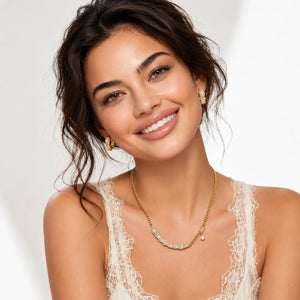 A smiling woman with dark hair models a delicate gold necklace with small clear stones and matching gold hoop earrings against a bright white background.