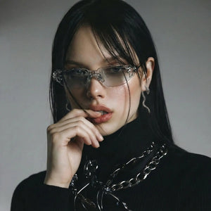 Person wearing glasses and a black outfit with a chain detail on a gray background