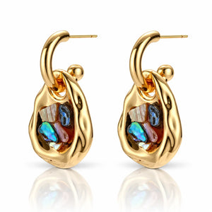 A pair of gold huggie hoop earrings with abstract organic-shaped gold bodies and colorful iridescent inlay stones, shown on a clean white background with a soft reflection below.