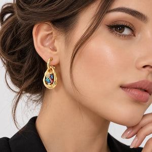 A close-up of a woman wearing a gold teardrop-shaped drop earring with colorful enamel details, set against a clean light gray background.