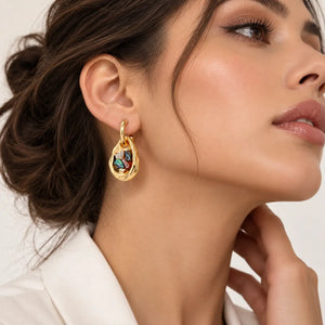 A close-up of a woman wearing a gold teardrop-shaped drop earring with a colorful enamel center, set against a clean light background.