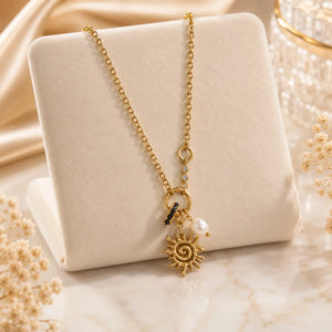 A gold chain necklace with a sun pendant, a pearl bead, and a small black stone charm, displayed on a cream-colored jewelry stand.