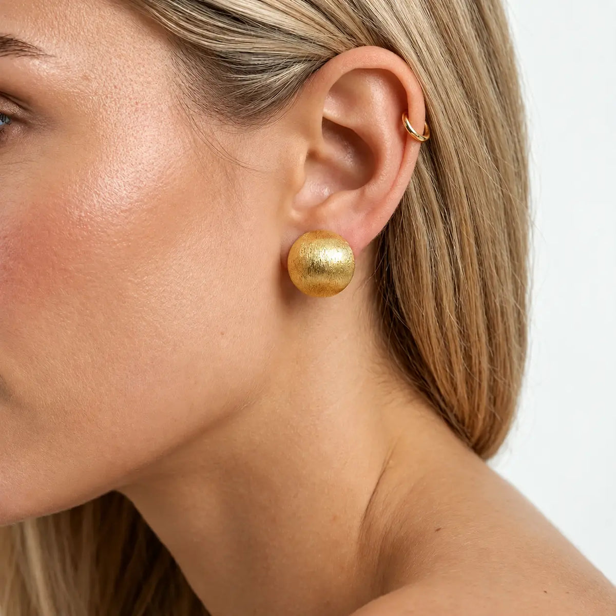 A close-up of a woman's ear wearing a textured gold spherical stud earring and a small gold hoop ear cuff, against a clean white background.