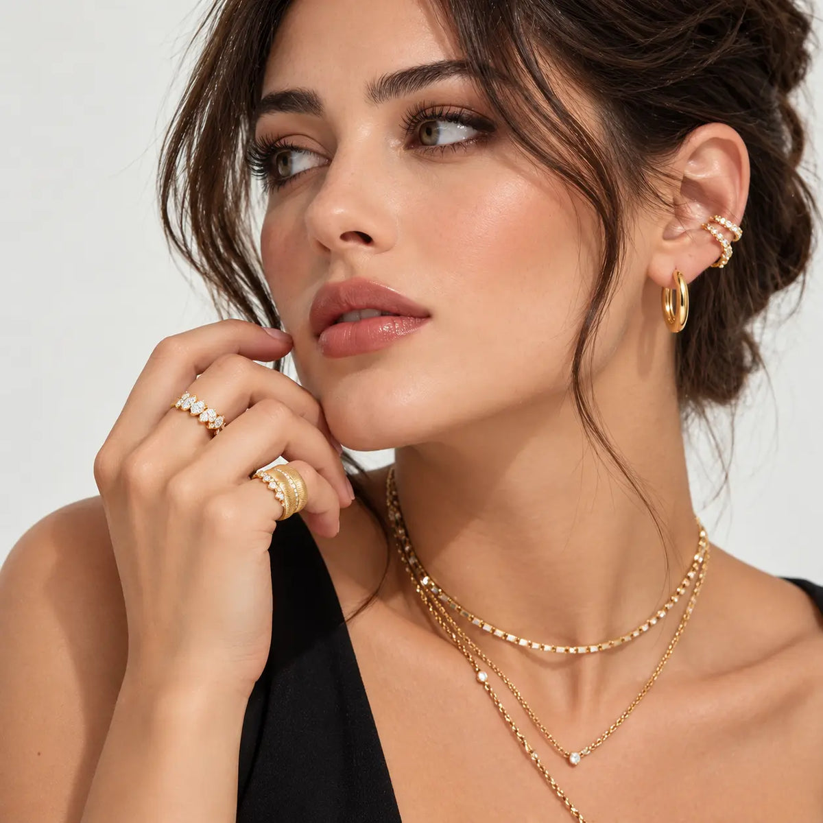 A close-up lifestyle shot of a woman wearing gold jewelry, including layered gold necklaces, rings, and hoop earrings, against a clean light gray background.