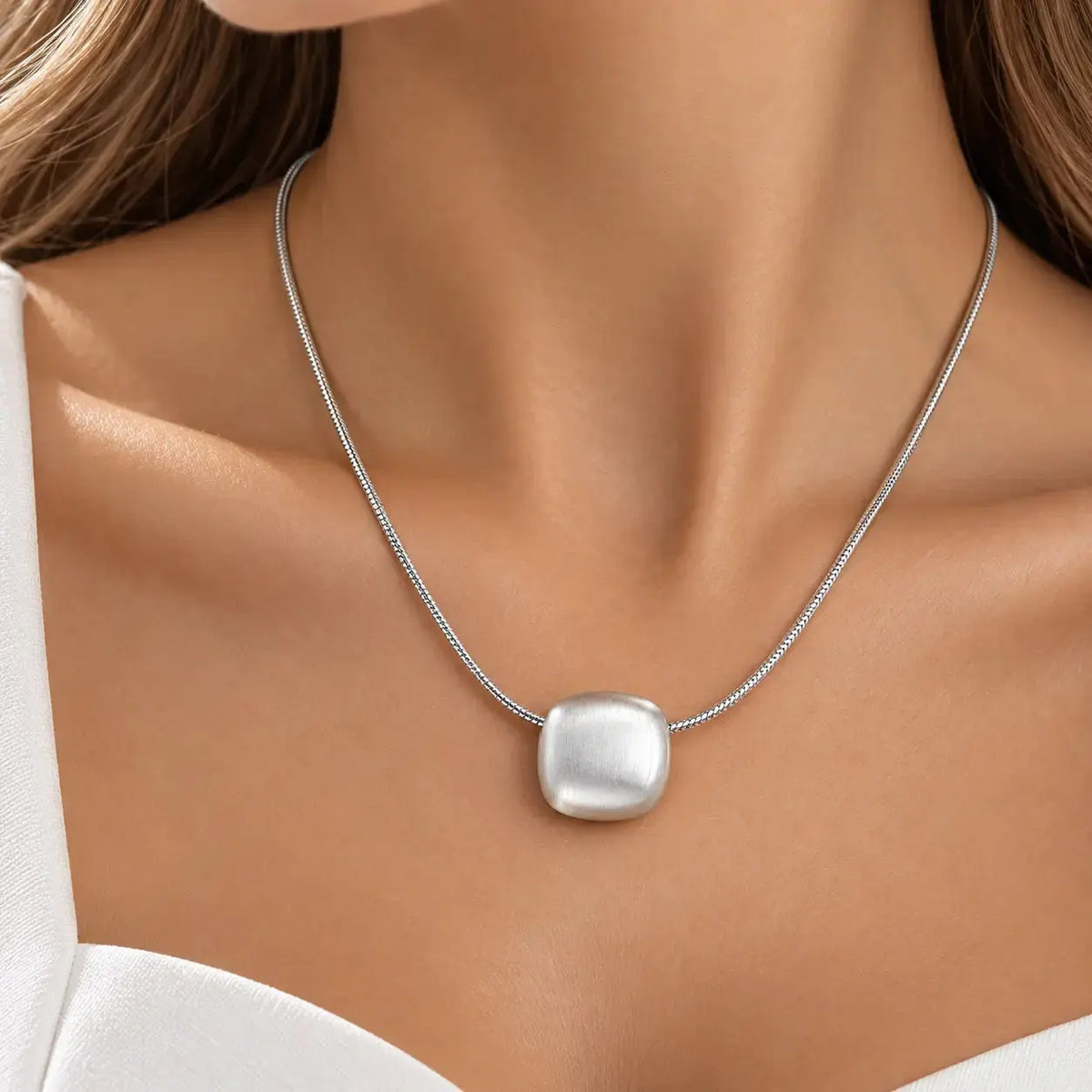 A close-up of a model's neck and chest wearing a silver snake chain necklace with a large, polished, square-shaped silver pendant.