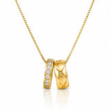 A gold pendant necklace featuring two interlocking bands: one with a row of clear stones and the other with an X-shaped cutout pattern, suspended on a gold chain against a white background.