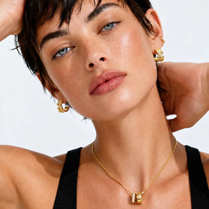 Close-up of a model wearing gold jewelry, including a delicate gold chain necklace with a small pendant and matching gold hoop earrings, against a clean white background.
