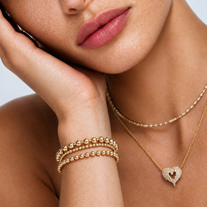 Close-up of a model wearing gold jewelry, including a beaded bracelet on the wrist, a delicate beaded necklace, and a gold heart pendant necklace with a heart-shaped crystal inlay.