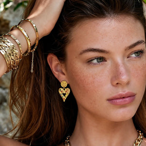 A close-up lifestyle shot of a woman with freckles wearing a gold heart-shaped dangle earring with a small red heart in the center, along with stacked gold bangles on her wrist.