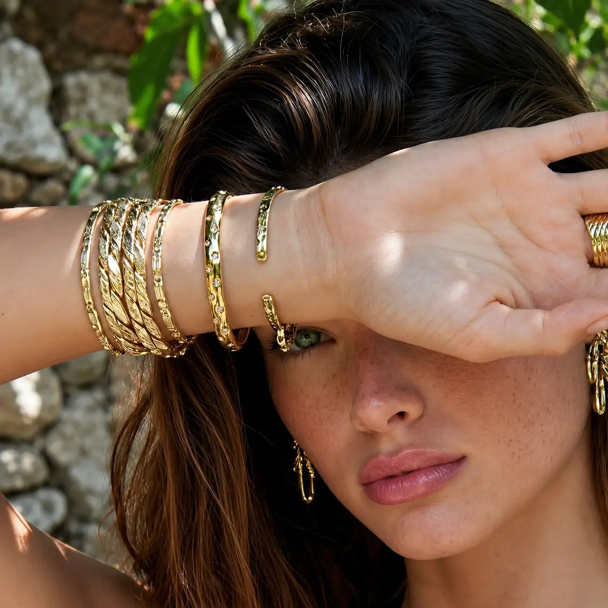 A close-up lifestyle shot of a woman wearing multiple gold-toned bangle bracelets and rings, with her hand covering part of her face. She is wearing gold hoop earrings, set against a blurred outdoor background of green foliage and a stone wall.