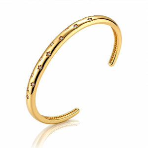A polished gold open cuff bracelet featuring a row of small clear gemstones set along the front, displayed on a clean white background with a soft reflection.