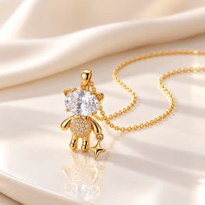 A gold teddy bear pendant necklace with a large clear heart-shaped crystal and a small star charm, displayed on a reflective white surface with draped satin fabric in the background.