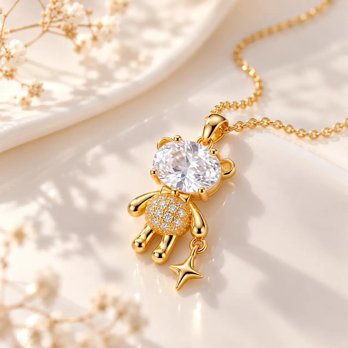 A gold teddy bear pendant necklace with a large clear round crystal for the face and small clear stones on the body, shown on a white surface with soft decorative flowers in the background.