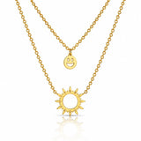 A double-layered gold necklace set featuring a delicate cable chain with a small smiley face disc pendant and a thicker link chain with a sunburst pendant, displayed on a clean white background with a soft reflection.