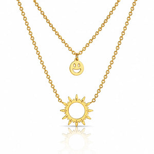 A double-layered gold necklace set featuring a delicate cable chain with a small smiley face disc pendant and a thicker link chain with a sunburst pendant, displayed on a clean white background with a soft reflection.