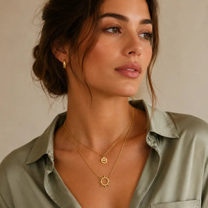 A close-up of a woman wearing layered gold necklaces, including a smiley face pendant and a sun pendant, along with a small gold hoop earring, paired with an olive green satin shirt against a neutral beige background.