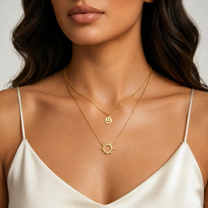 A close-up of a model wearing a double-layered gold necklace set. The top layer features a delicate chain with a small smiley face charm, and the bottom layer has a slightly thicker chain with a gold sun pendant.