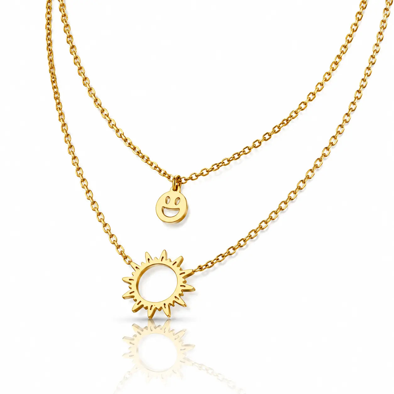 A double-layered gold necklace set on a clean white background with a soft reflection below. The top layer features a delicate cable chain with a small circular charm engraved with a smiley face, while the bottom layer has a slightly thicker curb chain with a larger sunburst pendant containing a smiley face detail.