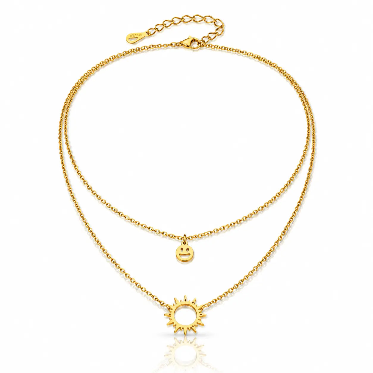 A double-layered gold necklace featuring a delicate chain with a small smiley face charm and a larger sun pendant, displayed on a clean white background with a subtle reflection.